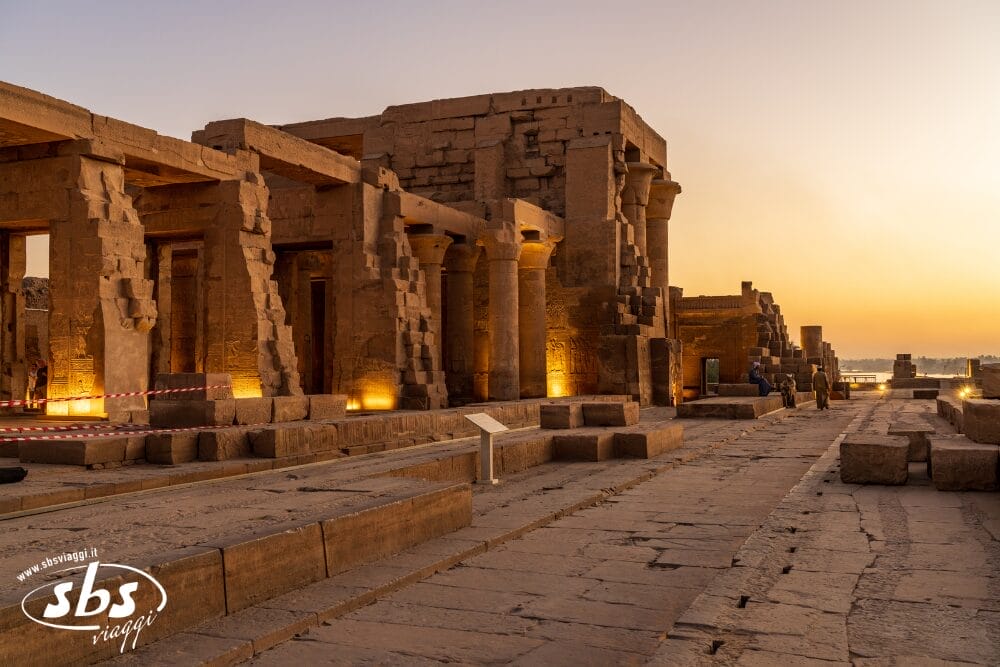 Antiche rovine in pietra con colonne al tramonto, illuminate dalle luci del pavimento. Sullo sfondo sono visibili poche persone. Il terreno è pavimentato con grandi lastre di pietra sotto un cielo dorato. Bozza automatica e logo "sbs viaggi" in basso a sinistra.