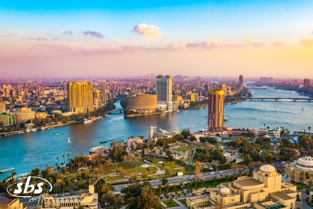 Veduta aerea del Cairo al tramonto, che mostra il fiume Nilo che si snoda attraverso la città con alti edifici, parchi verdi, ponti e un tocco di Bozza automatica sotto un cielo parzialmente nuvoloso.