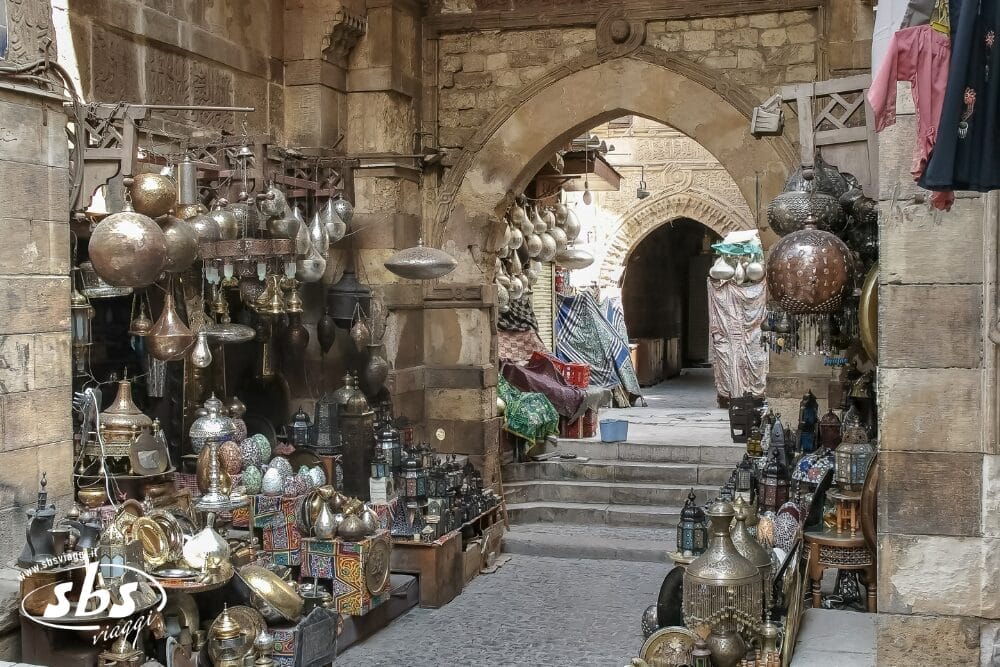 Stretto vicolo di un tradizionale mercato mediorientale con archi in pietra, negozi che espongono lampade di metallo ornate, lanterne e ceramiche automatiche Bozza su entrambi i lati e tessuti colorati sullo sfondo.