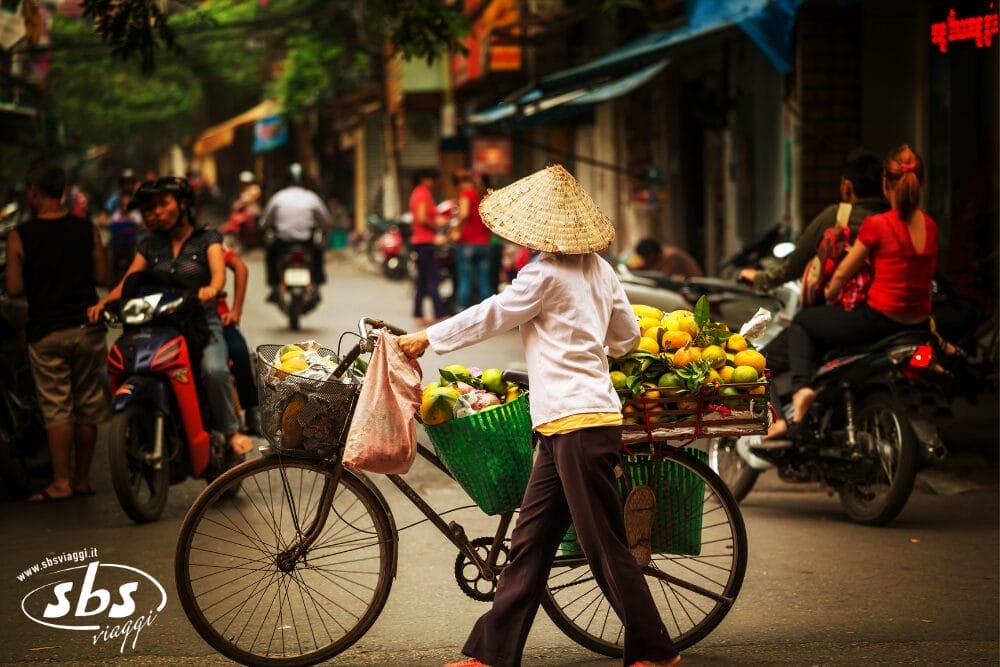 Un venditore ambulante con un cappello conico porta a spasso una bicicletta carica di cesti di frutta in una strada affollata e vivace, mentre persone e motociclette passano davanti alla scena animata come in un'istantanea di Bozza automatica.