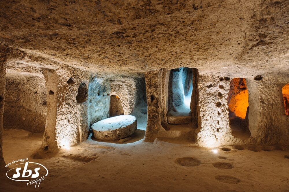 Una grotta sotterranea illuminata con pareti di pietra grezza, molteplici porte e una grande pietra rotonda al centro. Alcuni passaggi sono illuminati da luci arancioni e blu. Perfetto per il vostro Tour della Cappadocia. Il logo di SBS Viaggi è visibile nell'angolo in basso a sinistra.