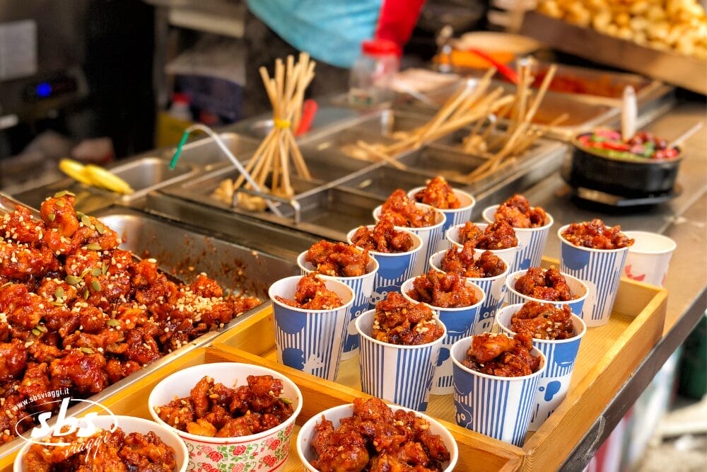Tazze riempite con pezzi di pollo fritto glassato sono disposte su un vassoio di legno in una bancarella di cibo, che mostra un allestimento automatico Bozza. Spiedini e vassoi con vari ingredienti contribuiscono alla vivace scena del mercato sullo sfondo.