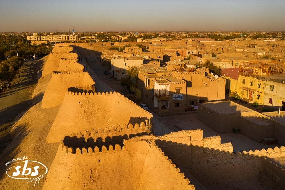 Vista aerea delle antiche mura di Khiva che proiettano lunghe ombre sugli edifici sottostanti, con un mix di strutture storiche e moderne sotto un cielo limpido, che cattura il fascino della città vecchia attraverso una prospettiva automatica di Bozza.