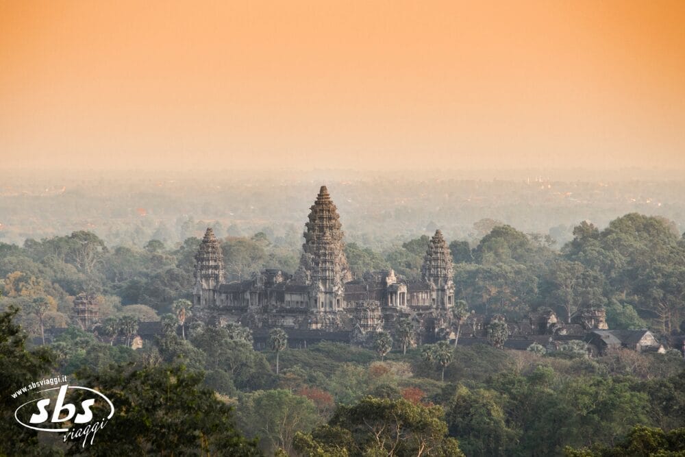 L'antico tempio di Angkor Wat si erge tra una fitta foresta verde, con un cielo arancione all'alba o al tramonto sullo sfondo. Scoprite questa meraviglia durante un tour Cambogia, dove le torri del tempio si ergono sopra la vegetazione lussureggiante.
