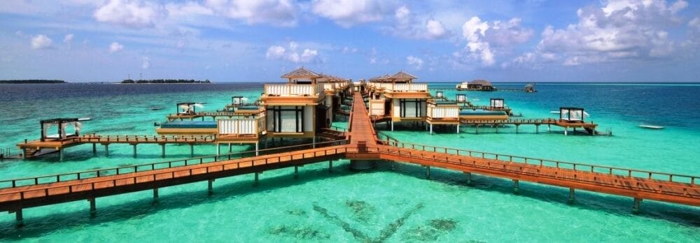 I bungalow overwater, collegati da passerelle di legno, si trovano sopra l'acqua limpida e turchese, con capanne sparse che si estendono nell'oceano sotto un cielo parzialmente nuvoloso, creando una scena serena che ricorda una Bozza automatica.