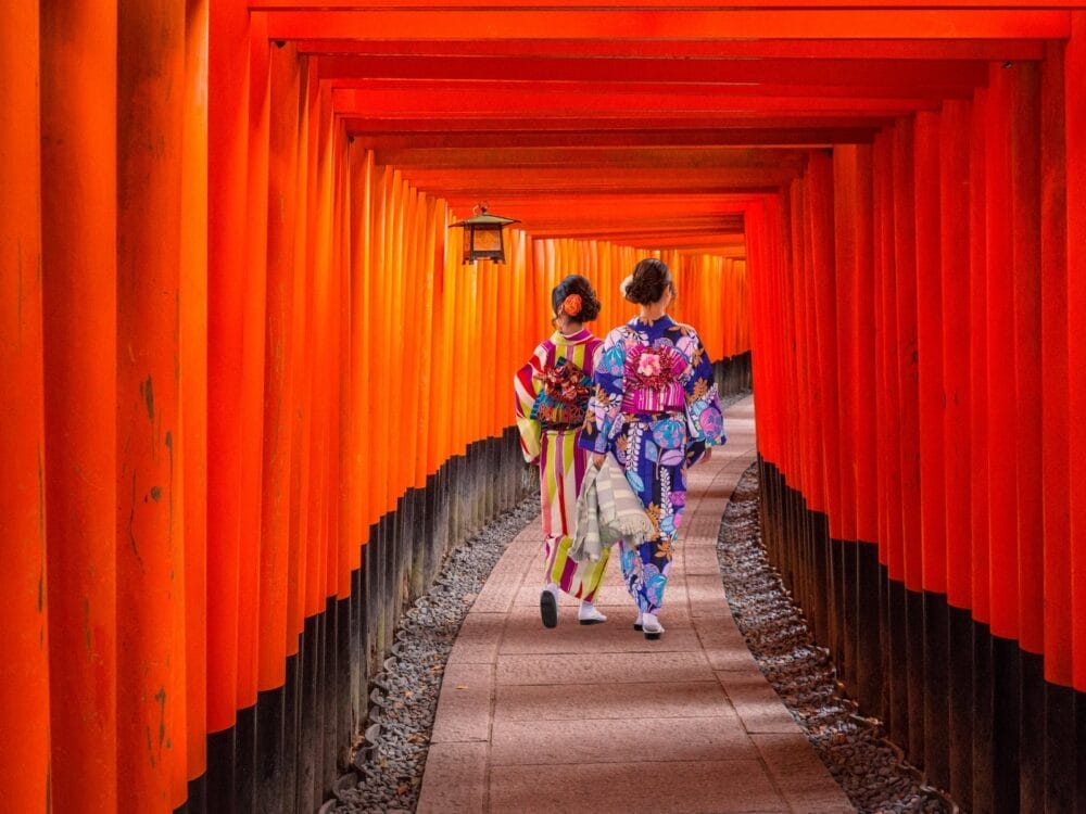 Due persone che indossano coloratissimi kimono tradizionali giapponesi attraversano un tunnel di porte torii di colore rosso vivo presso un santuario, con lanterne appese sopra il sentiero di pietra lastricato.