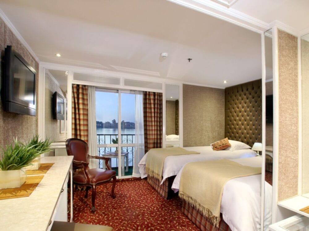 Una camera d'albergo con due letti, testata imbottita, moquette rossa, scrivania con pianta, sedia in pelle, TV a muro, grandi specchi e balcone con tende che si affacciano sull'acqua: un rifugio perfetto prima dell'avventura sulla Crociera Nilo.