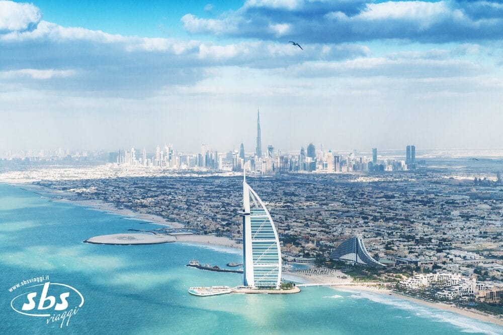 Vista aerea della costa di Dubai con l'hotel Burj Al Arab a forma di vela, le acque turchesi, le spiagge sabbiose e lo skyline della città con il Burj Khalifa, una vista iconica di qualsiasi tour di Dubai, sotto un cielo parzialmente nuvoloso.
