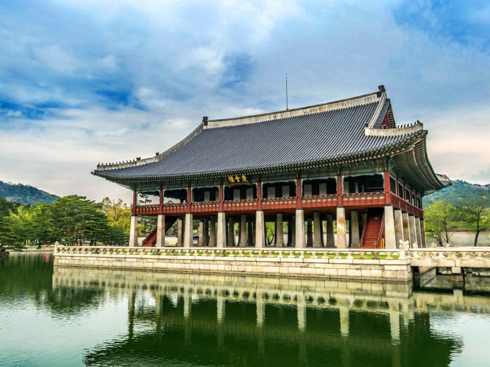 Edificio tradizionale coreano con tetto di tegole, pilastri rossi e base in pietra bianca, riflesso in un tranquillo stagno: una scena iconica della Corea del Sud, che evoca la serenità e la bellezza senza tempo del sogno coreano.