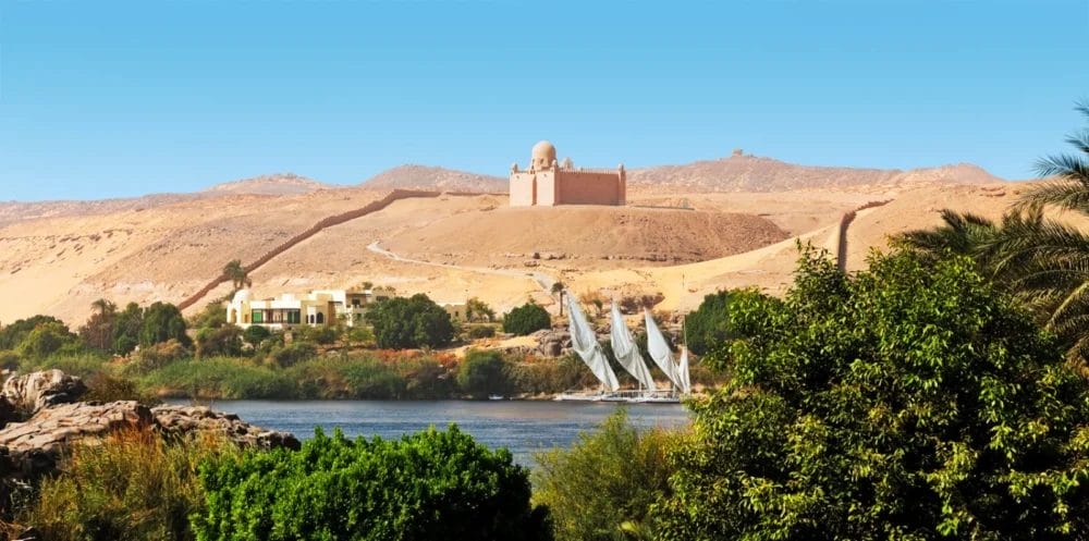 Una vista panoramica del fiume Nilo vicino a Luxor con alberi verdi in primo piano, barche a vela bianche della Crociera Ramses sull'acqua e un edificio a cupola in cima a una collina sabbiosa sotto un cielo blu chiaro sullo sfondo.