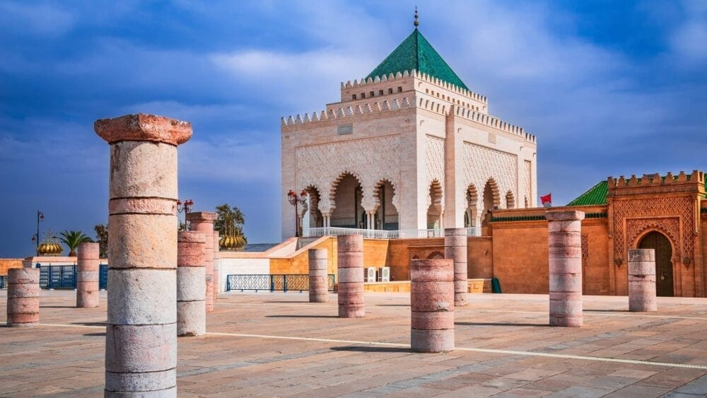 Un mausoleo in marmo bianco con un tetto di tegole verdi e archi ornati si erge dietro antiche colonne di pietra sotto un cielo azzurro a Rabat, in Marocco: una tappa obbligata del vostro Gran Tour del Marocco o di qualsiasi altro indimenticabile Tour Marocco.