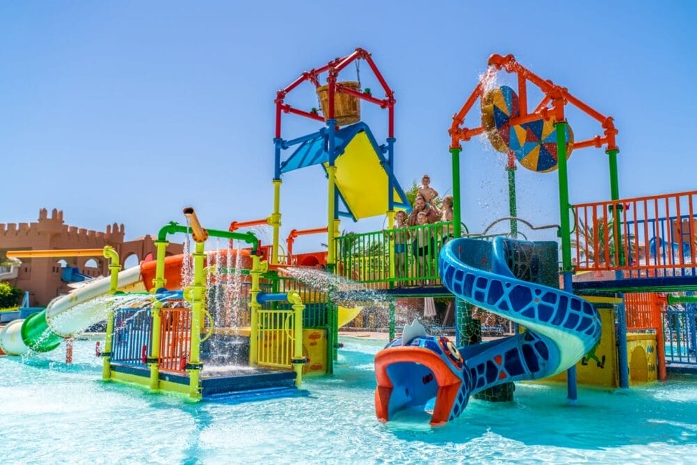 I bambini giocano e si divertono nel soleggiato parco acquatico all'aperto dell'Akassia Swiss Beach Resort, con scivoli dai colori vivaci, strutture per l'arrampicata e secchi ribaltabili che spruzzano acqua sotto un cielo azzurro e limpido.