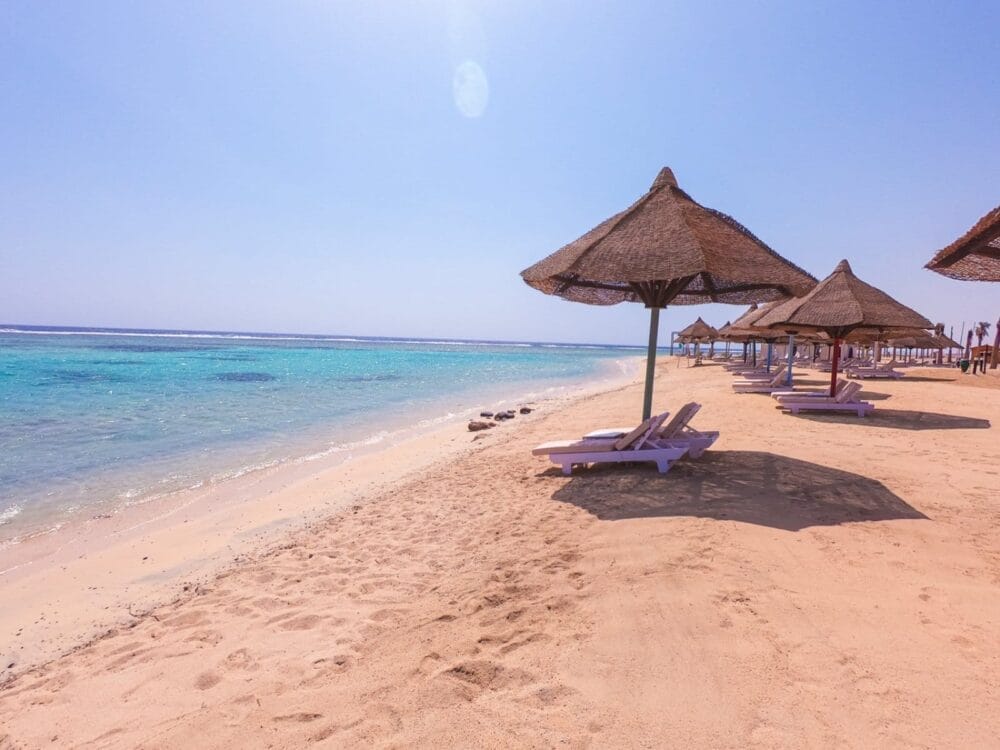 Spiaggia sabbiosa con ombrelloni di paglia e sedie a sdraio vuote che si affacciano sull'acqua azzurra dell'oceano sotto un cielo luminoso e soleggiato: una fuga degna di Alessandro Magno.