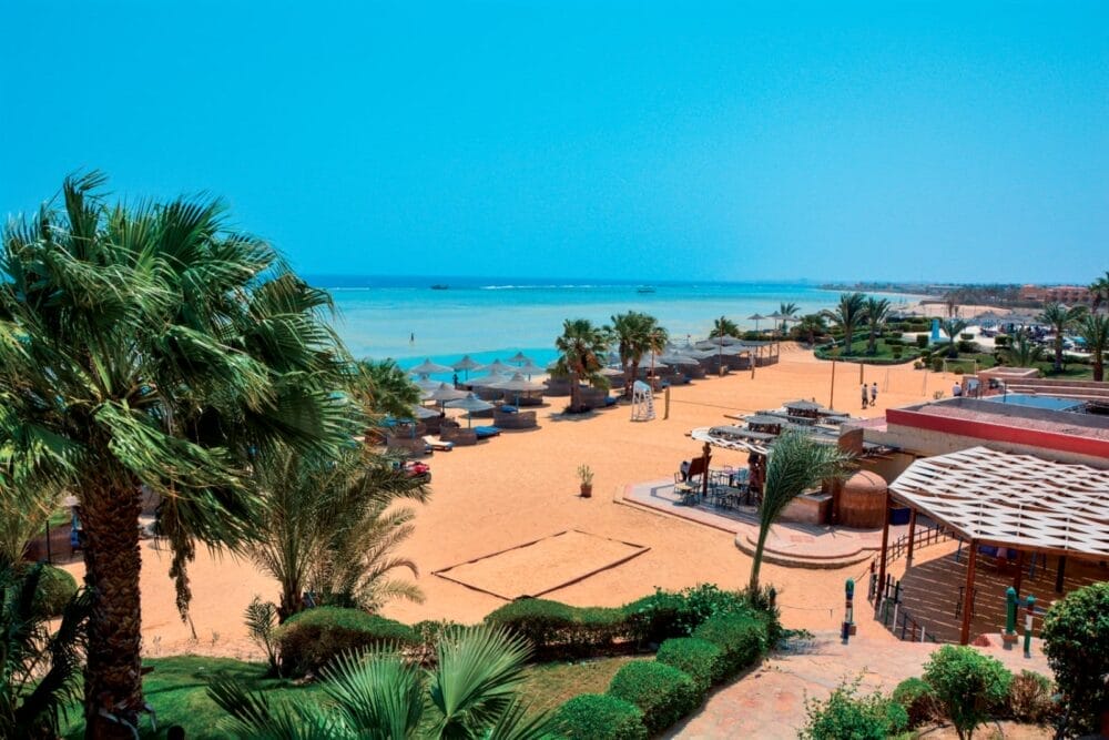 Una spiaggia soleggiata con sabbia dorata, palme e lettini all'ombra degli ombrelloni, vicino a un mare azzurro e calmo. Gli ospiti del Blue Reef Ciaoclub passeggiano lungo la riva, mentre i sentieri paesaggistici conducono alle strutture sulla spiaggia.