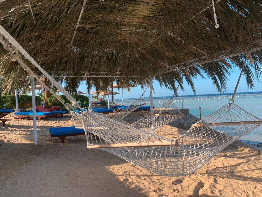Due amache di corda sono appese sotto un tetto di paglia sulla spiaggia sabbiosa di Blue Reef, con sedie a sdraio blu nelle vicinanze e l'acqua calma dell'oceano sullo sfondo. L'ambiente è tranquillo e invitante, perfetto per rilassarsi in stile Ciaoclub in riva al mare.