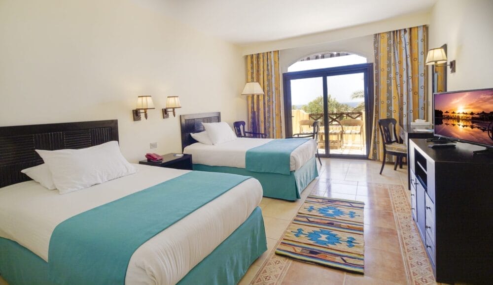 Una camera d'albergo dell'Elphistone Resort è dotata di due letti matrimoniali con tappetini color verde acqua, una TV su un comò, una scrivania e una sedia, tappeti a fantasia e ampie finestre con tende gialle che si aprono su un balcone con una vista mozzafiato.