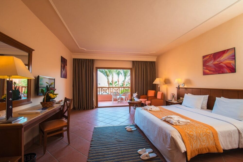 Un'accogliente camera d'albergo al Charmillion Sea Life Resort con un grande letto, accenti arancioni, scrivania, specchio, due sedie e lampade. Godetevi un balcone con vista sulle palme in questo resort sulla spiaggia di Sharm El Sheikh. Pantofole e asciugamani sono disposti in modo ordinato per il vostro comfort.