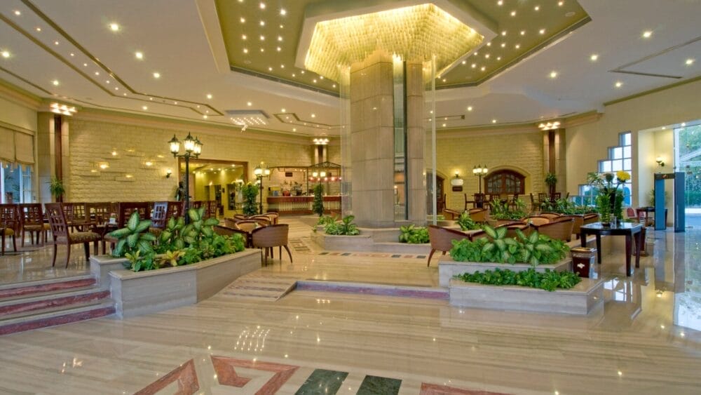 Ampia hall dell'hotel Dreams Beach con pavimenti lucidi, pilastro centrale, illuminazione moderna, piante da interno, sedie e tavoli in legno e banco della reception sullo sfondo. Le grandi finestre inondano lo spazio di luce naturale.