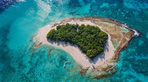 Vista aerea di una piccola isola verde e lussureggiante circondata da acque turchesi e spiagge di sabbia bianca, con la barriera corallina visibile sotto la superficie: una scena idilliaca che ricorda un lussuoso resort delle Maldive come il Ja Manafaru Resort.