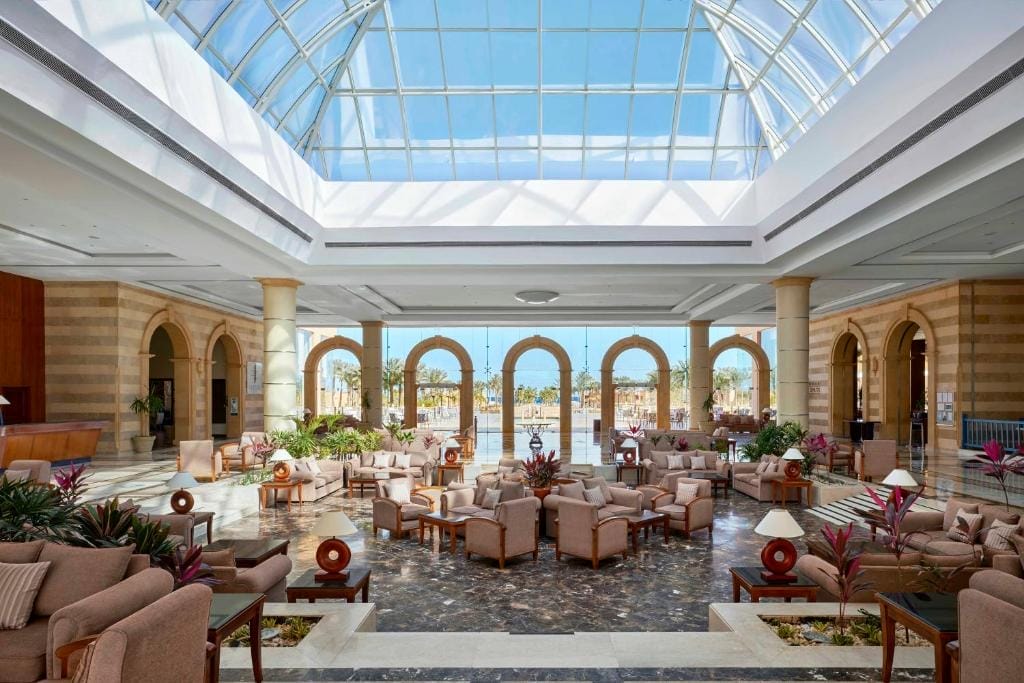 La spaziosa hall del Jaz Grand Marsa presenta un soffitto a cupola in vetro, finestre ad arco, pareti beige, pavimenti in marmo e aree salotto con poltrone e tavolini beige. La luce del sole riempie la stanza, mettendo in risalto le piante in vaso e l'arredamento moderno.