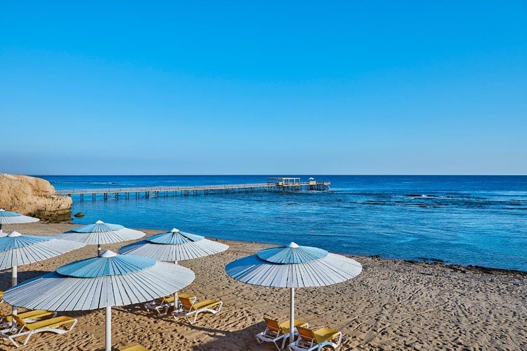 Una spiaggia sabbiosa con ombrelloni bianchi e sedie a sdraio gialle si affaccia sulle calme acque blu del Mar Rosso. In lontananza, un lungo molo di legno si estende sotto un cielo limpido e luminoso al Jaz Grand Marsa di Marsa Alam.