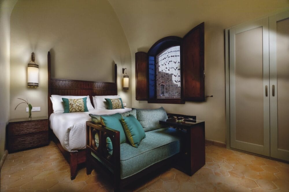 Accogliente camera d'albergo del Movenpick Resort El Quseir con un letto matrimoniale, cuscini accesi verde acqua, un piccolo divano ai piedi, mobili in legno, una finestra con persiane aperte e un'illuminazione calda.