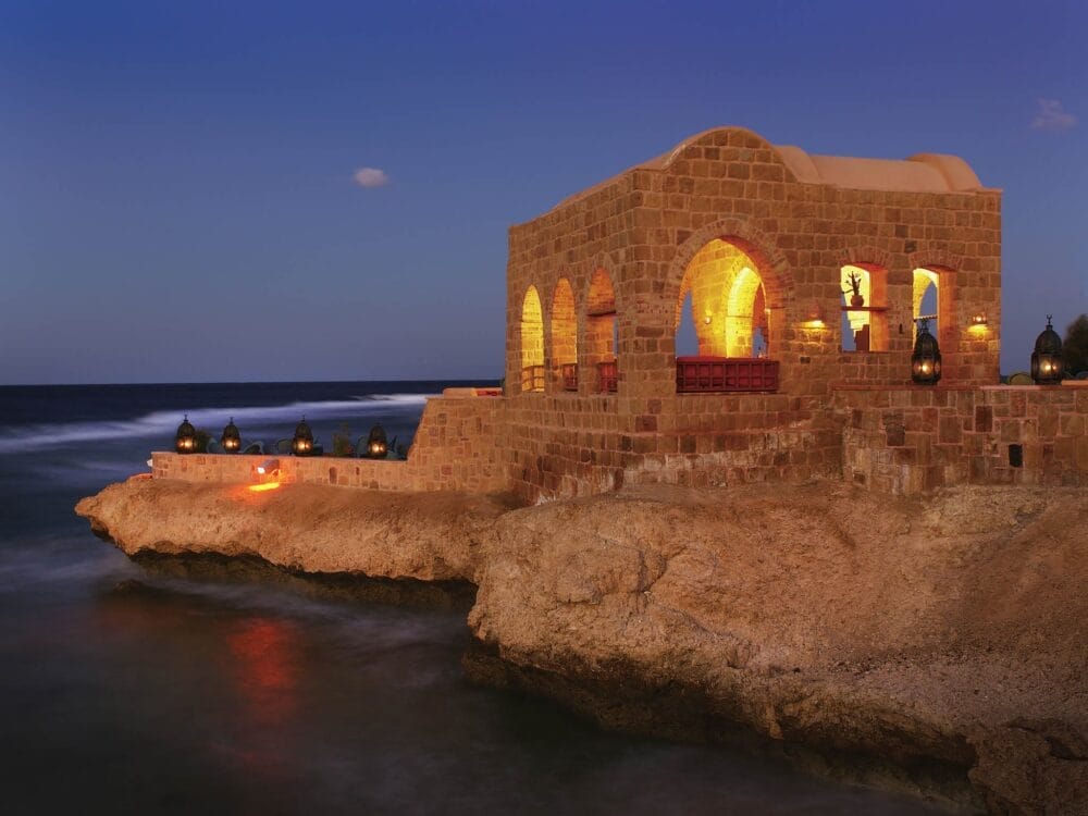 Un edificio in pietra con finestre ad arco che risplendono di luce calda si trova su una costa rocciosa in riva al mare al tramonto, con lanterne decorative disposte lungo il bordo dell'incantevole Movenpick Resort El Quseir.