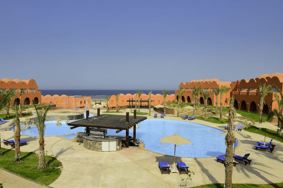 Il Novotel Marsa Alam è un resort sul Mar Rosso con edifici ad arco arancione che circondano una piscina in stile laguna e un bar ombreggiato. Sedie a sdraio blu, palme e vista sul mare sotto un cielo limpido creano la perfetta fuga dall'hotel di Marsa Alam.