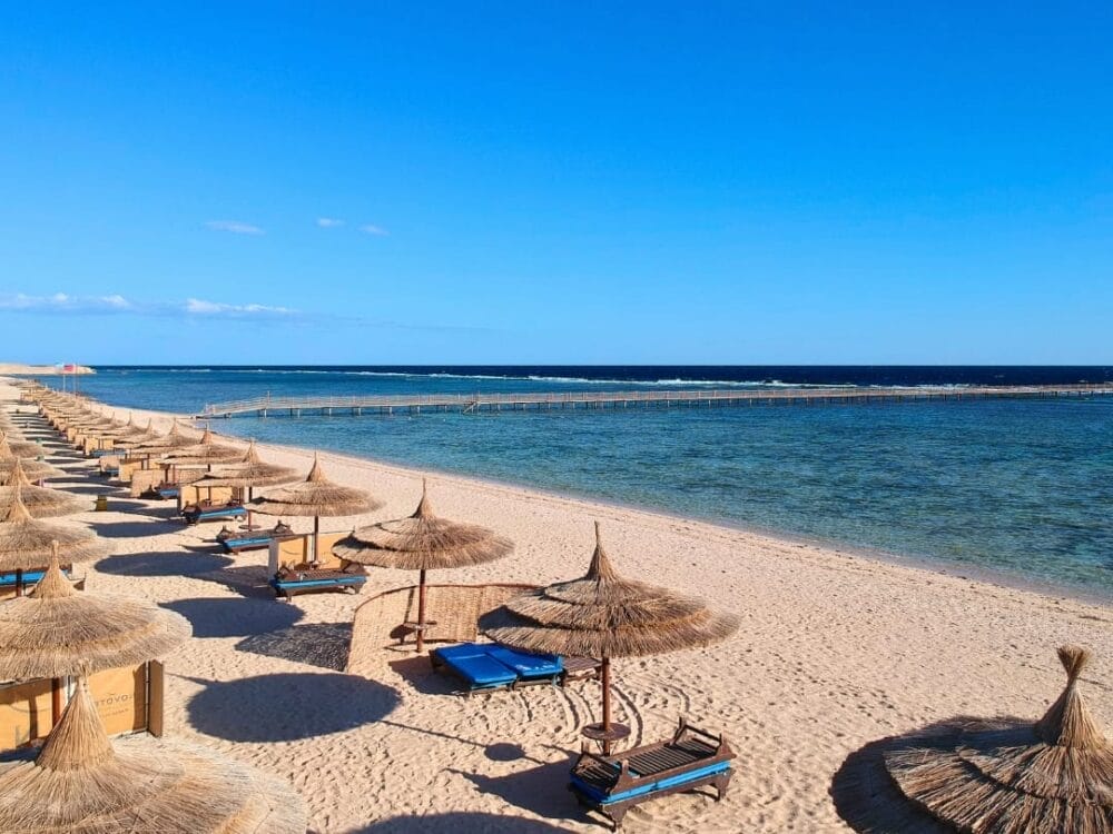 Una spiaggia sabbiosa con file di ombrelloni e sedie a sdraio si affaccia sul mare calmo e cristallino del Novotel Marsa Alam, un resort sul Mar Rosso. Sullo sfondo, sotto un cielo luminoso, un molo di legno si estende sull'acqua.