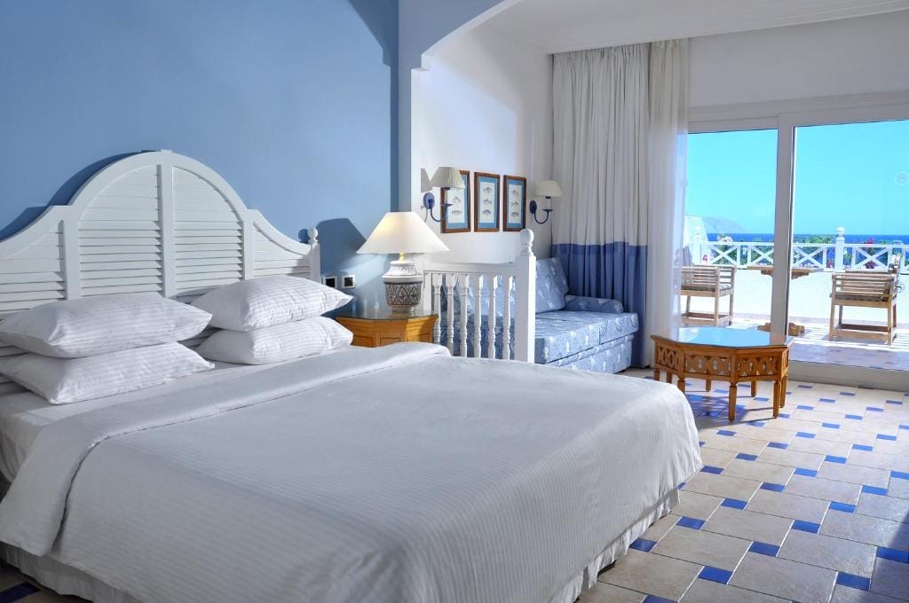 Una luminosa camera d'albergo dello Sheraton Sharm Resort con un grande letto, decorazioni blu e bianche, un divano, un pavimento piastrellato, un tavolino con lampada e porte di vetro che conducono a un balcone con sedie e una splendida vista sulla spiaggia di Sharm El Sheikh.