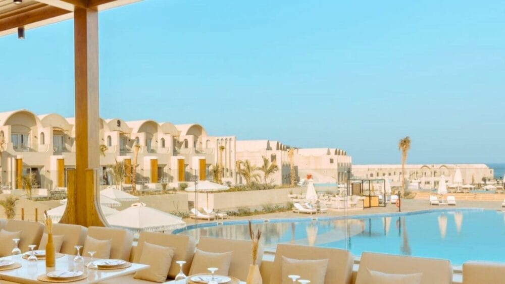 I posti a sedere del ristorante all'aperto del Sunrise Anjum Resort si affacciano su una grande piscina, con sedie a sdraio bianche, ombrelloni e moderni edifici bianchi sotto un cielo azzurro e limpido.