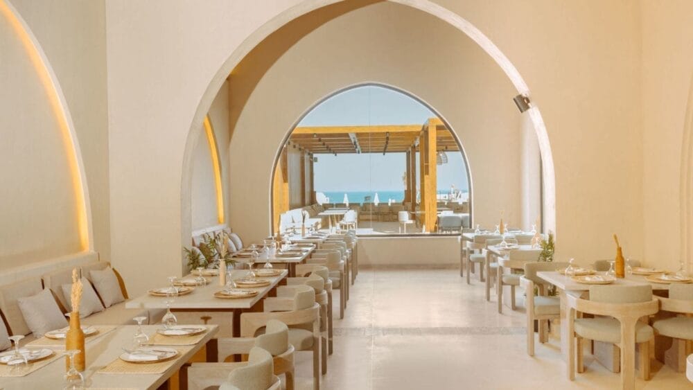 Elegante interno del ristorante Casa Sand, con porte ad arco, pareti dai toni neutri, tavoli in legno per mangiare, sedie imbottite e vista sul patio esterno e sul mare sullo sfondo.