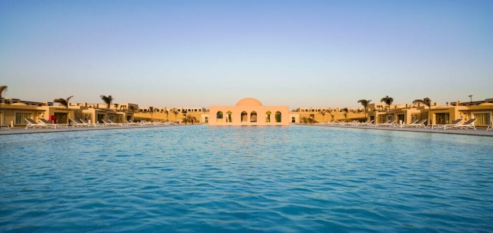 Grande e serena piscina all'aperto con acqua cristallina all'Oasi Valtur Reef, circondata da ville moderne e palme sotto un cielo limpido; un edificio centrale a cupola bianca con archi si trova all'estremità della piscina.