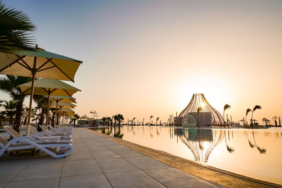 File di sedie a sdraio bianche e ombrelloni gialli costeggiano la piscina Reef Oasis al tramonto, con palme e una cupola a struttura aperta unica nel suo genere che si riflette nell'acqua calma. Il cielo è limpido e dorato.