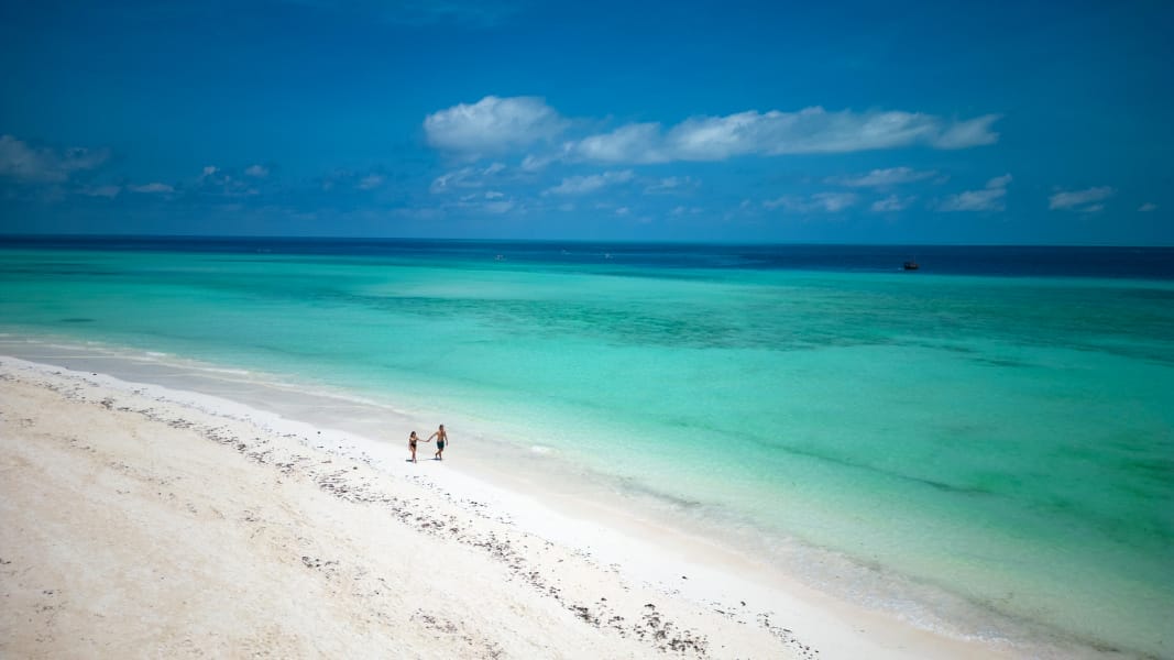 Una coppia cammina mano nella mano lungo la spiaggia incontaminata di sabbia bianca del Mora Resort, accanto a un'acqua limpida e turchese, sotto un cielo azzurro con nuvole sparse.