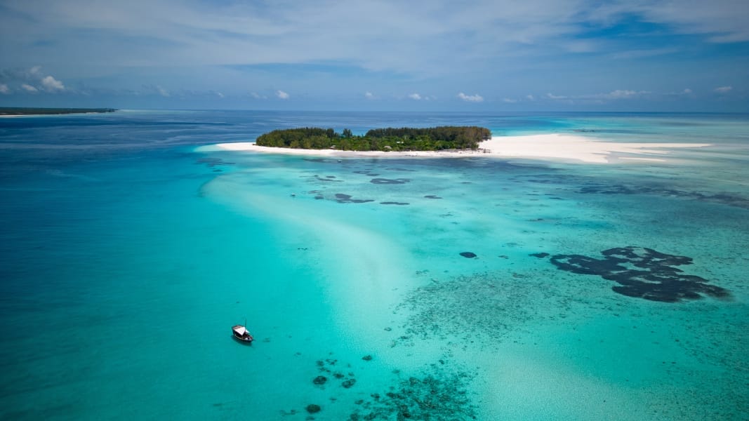Una piccola isola lussureggiante circondata da acque oceaniche turchesi e blu intenso, con spiagge bianche e sabbiose vicino a un resort di Zanzibar. Una barca galleggia nelle vicinanze sotto un cielo parzialmente nuvoloso e le barriere coralline sono visibili sotto l'acqua limpida.