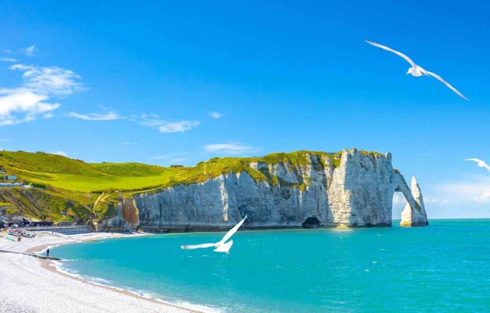 Le bianche scogliere di gesso con un arco naturale si ergono sul mare turchese di Etretat, in Normandia, sotto un cielo blu brillante. Una spiaggia di ciottoli fiancheggia la riva, con le colline verdi sopra e i gabbiani che volano in alto: l'ideale per un tour panoramico.