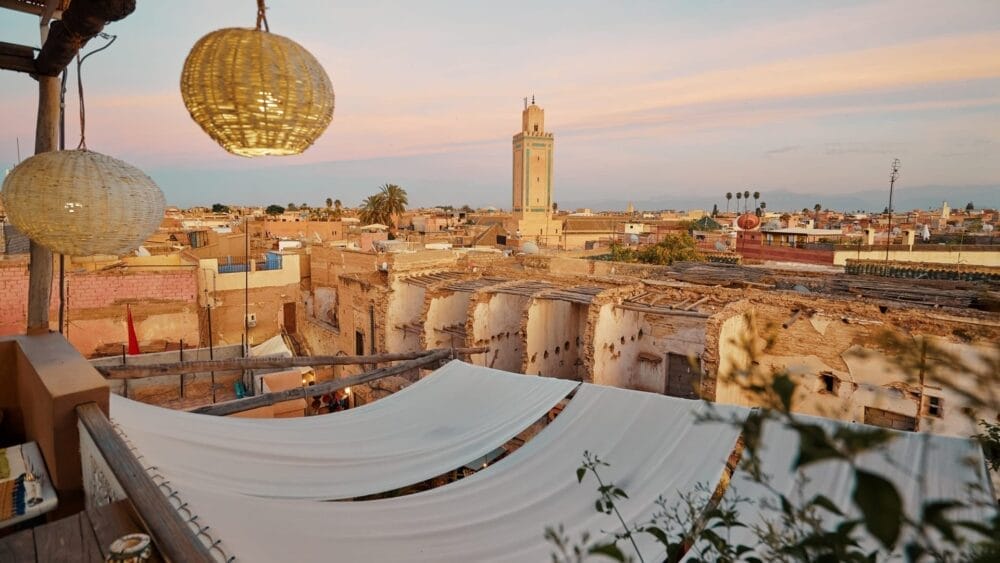 Una terrazza sul tetto con lanterne sospese si affaccia sulla città vecchia di Marrakech al tramonto, invitandovi a vivere la magia di un Gran Tour del Marocco, con edifici tradizionali e l'alto minareto di una moschea sotto un cielo pastello.