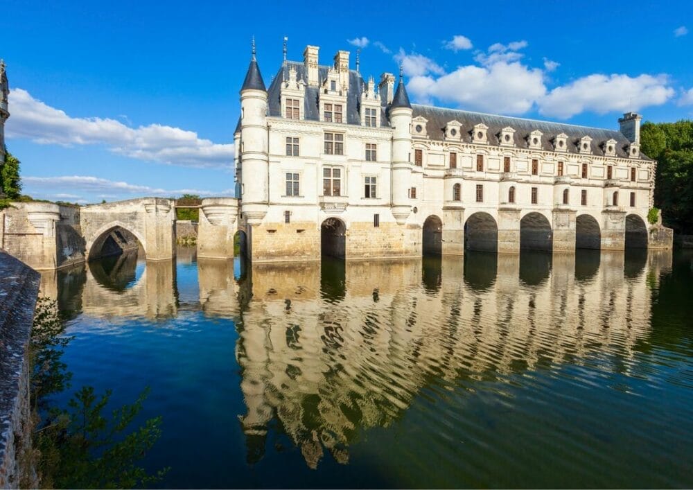 Un castello in pietra bianca con ponti ad arco si trova su un fiume tranquillo, con la sua Bozza automatica che si riflette perfettamente nell'acqua sottostante. Il cielo è azzurro con nuvole sparse e una vegetazione lussureggiante circonda l'edificio storico.