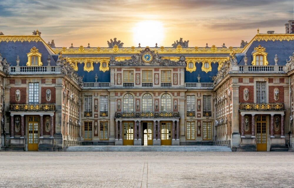 Il sole tramonta dietro la facciata ornata di Versailles, vicino a Parigi, mettendo in risalto le sue decorazioni dorate, le finestre simmetriche e l'architettura grandiosa. In primo piano, l'ampio cortile in pietra è vuoto.