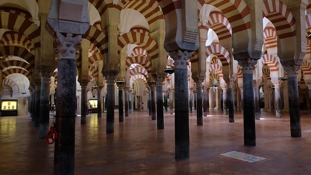 File di colonne in pietra decorate con archi a righe bianche e rosse, parte della Grande Moschea di Córdoba, in Spagna. L'ampia sala è caratterizzata da un pavimento piastrellato, una luce naturale soffusa e un senso di tranquillità, quasi come una perfetta bozza automatica.