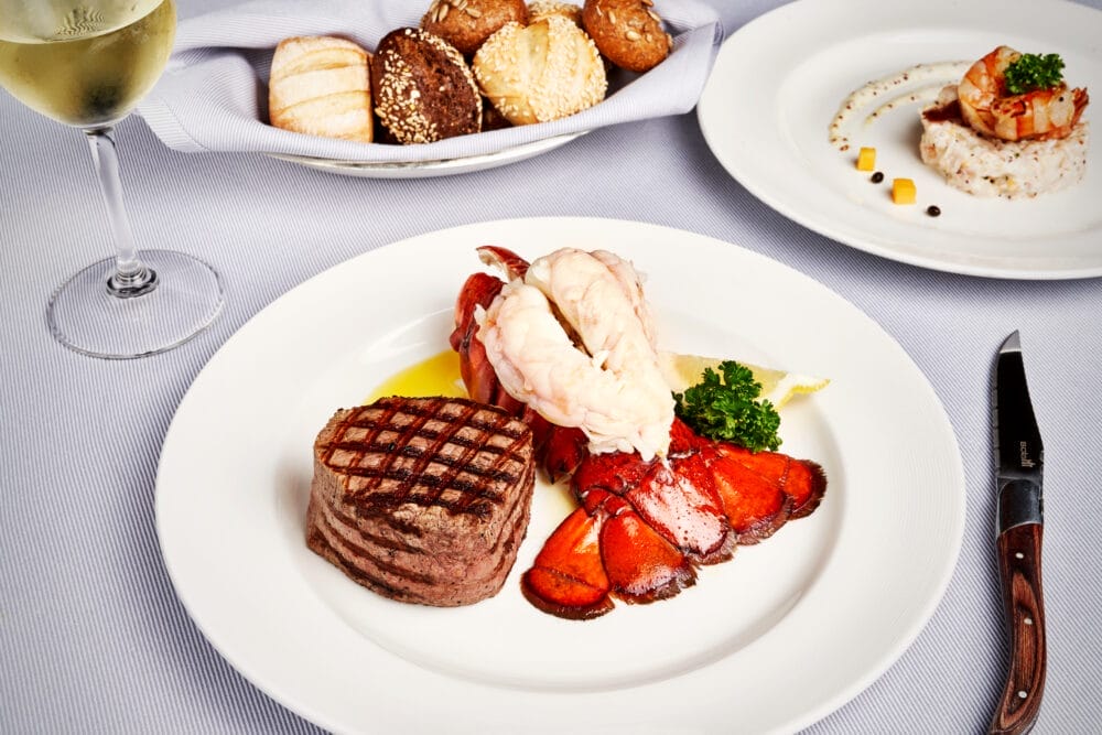 Un piatto di surf and turf con filet mignon alla griglia e coda di aragosta, guarnito con prezzemolo e spicchi di limone. Bozza automatica espone panini, un antipasto di gamberi, vino bianco e un coltello da bistecca su una tovaglia bianca.