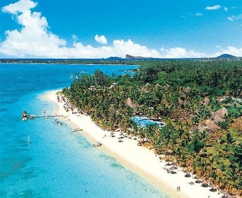 Veduta aerea del Trou Aux Biches Beachcomber Resort, con acque limpide e turchesi, sabbia bianca e palme verdi. Le persone si rilassano sulla spiaggia e nuotano sotto un cielo blu brillante con nuvole sparse.