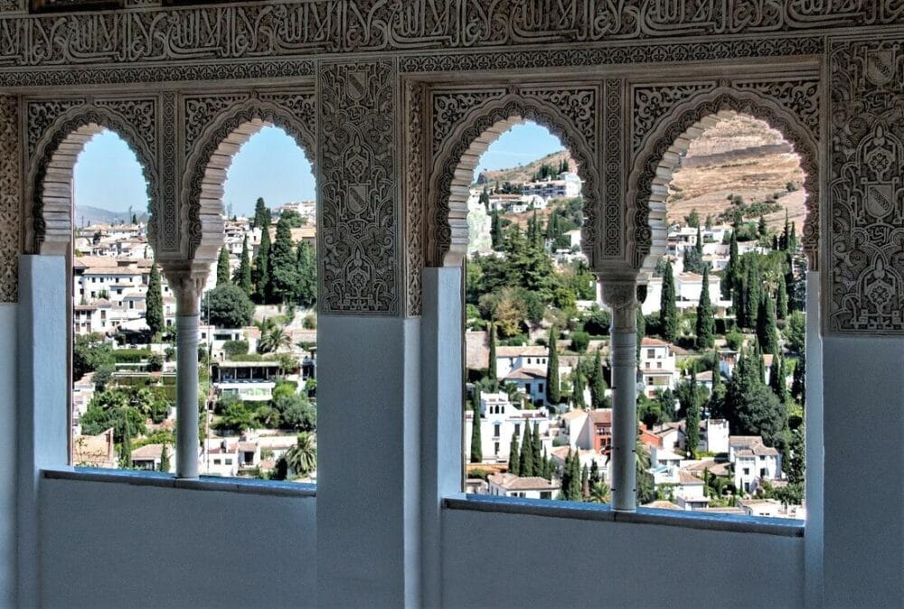 La vista di una città con edifici bianchi e verde, vista attraverso tre finestre ad arco con intagli intricati, evoca la ricca storia dell'Andalusia moresca - perfetta per il vostro Gran Tour Andalusia durante Capodanno 2025.