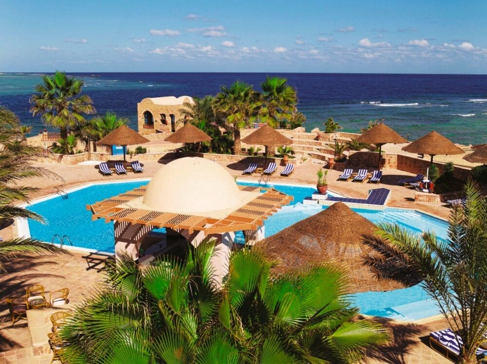 La lussuosa piscina del Mövenpick Resort El Quseir è caratterizzata da ombrelloni di paglia e sedie a sdraio che si affacciano sull'oceano blu. Palme e strutture in pietra circondano la piscina sotto un cielo soleggiato con nuvole sparse.