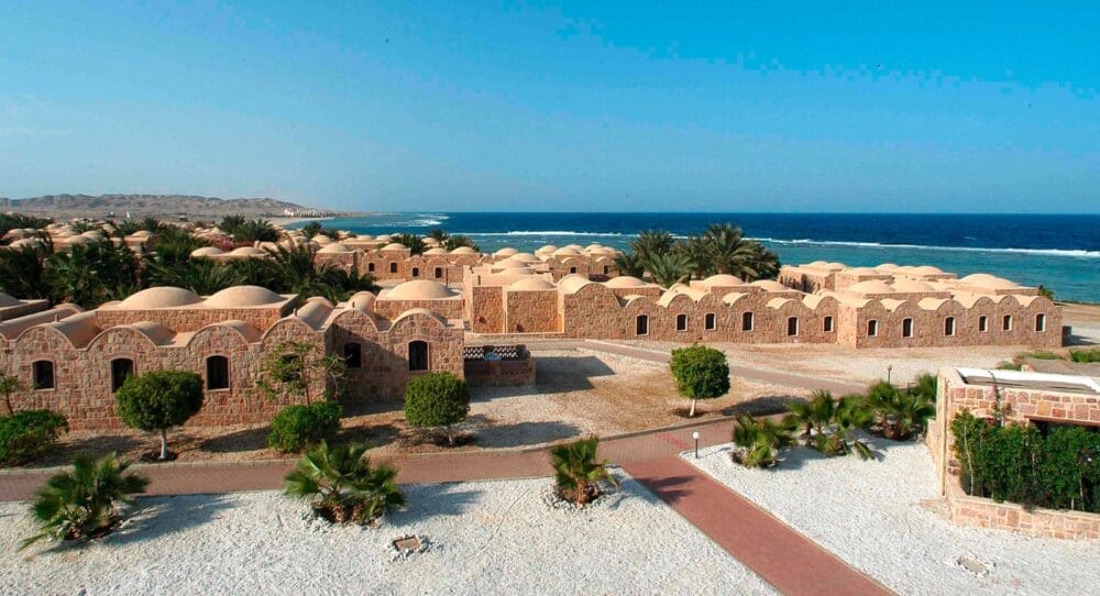 Un resort costiero con edifici in pietra dal tetto a cupola, circondato da palme e sentieri sabbiosi, il Mövenpick Resort El Quseir si affaccia su un mare blu sotto un cielo limpido.