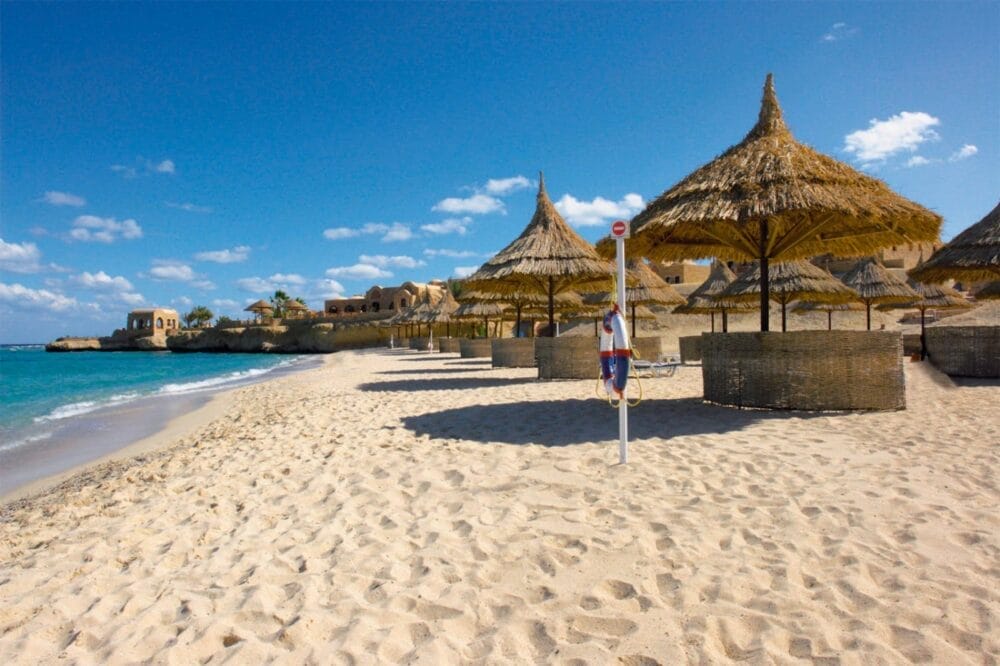 Una spiaggia sabbiosa con ombrelloni di paglia e ombrelloni di vimini, acqua turchese e cielo azzurro si estende vicino a un cartello. In lontananza si intravedono gli edifici che fanno parte del tranquillo Mövenpick Resort El Quseir, un resort iconico di El Quseir.