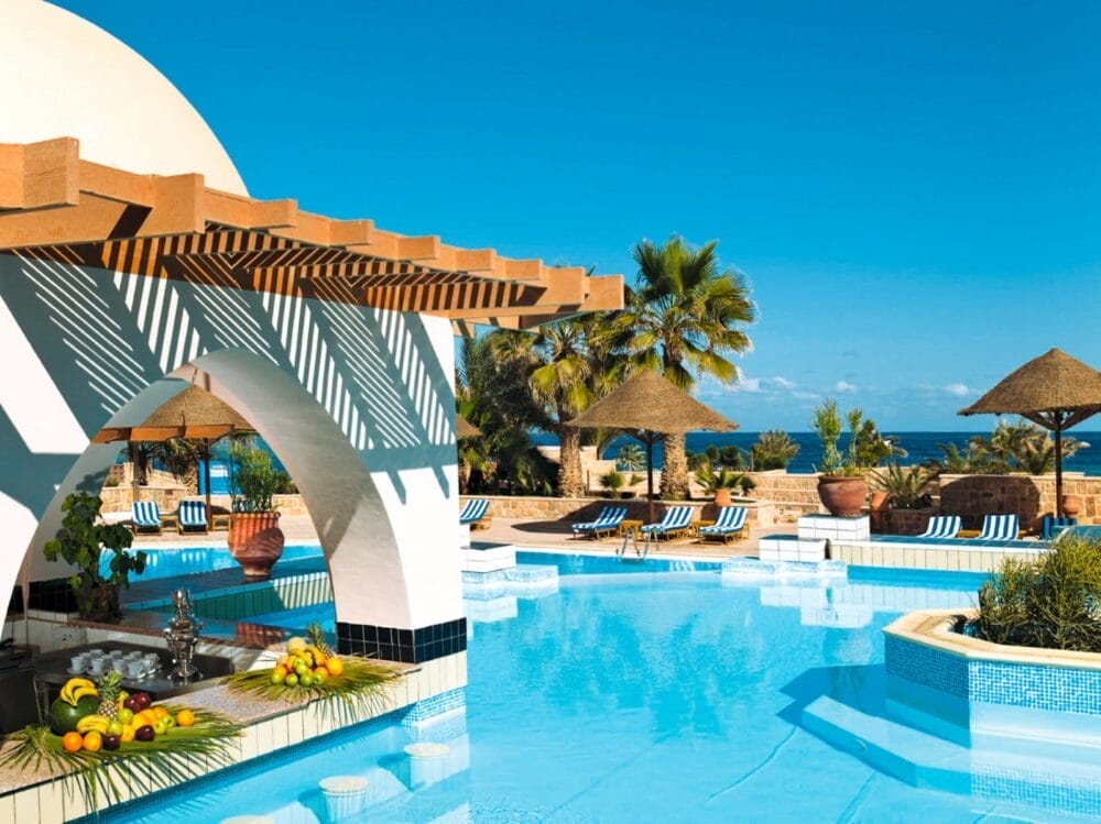 La piscina del Mövenpick Resort El Quseir, luminosa e soleggiata, è dotata di un bar ombreggiato da un pergolato e ornato di frutta fresca. Palme e sedie a sdraio a righe circondano la piscina, con lo splendido Mar Rosso sullo sfondo.