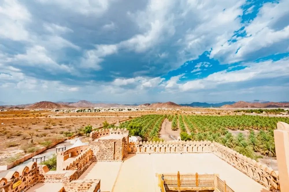 Una vista panoramica da una fortezza in pietra si affaccia su verdeggianti palmeti da dattero, terreni aridi, montagne lontane e un cielo blu parzialmente nuvoloso: una Bozza automatica che ispira i contrasti della natura.
