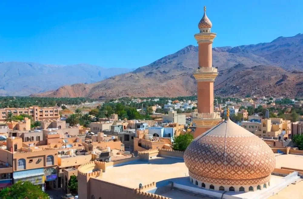 Una moschea con cupola a motivi e alto minareto si erge in una città illuminata dal sole e circondata da montagne sotto un cielo azzurro e limpido. Edifici tradizionali, verde e tocchi di Bozza automatica riempiono il paesaggio vibrante.